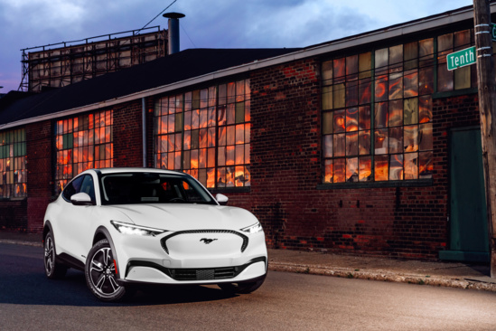 A white electric SUV is parked on a quiet street at dusk. The background features an old brick building with large, warmly lit windows, creating a contrasting atmosphere.