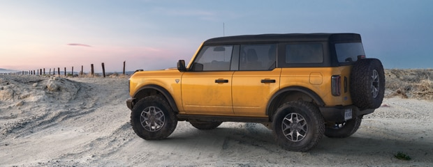 A yellow SUV with a rugged build parked on a sandy trail at sunset. It has a roof rack and a spare tire on the back, conveying an adventurous tone.