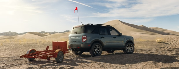 SUV with a red flag towing a small trailer in a sandy desert landscape, surrounded by dunes under a clear blue sky, conveying adventure and exploration.