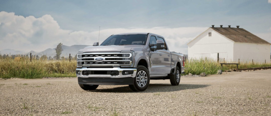 Silver pickup truck parked on a gravel road beside a grassy field and white barn. The sky is partly cloudy, creating a serene, rural atmosphere.