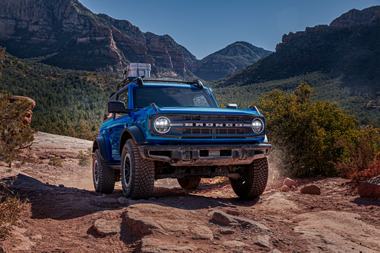 Blue SUV traverses rocky terrain in a scenic mountain landscape, emphasizing ruggedness and adventure. Bright sunlight highlights the vehicle’s front.