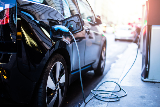 A black electric car is parked on a city street, plugged into a charging station. Sunlight reflects off the car's sleek surface, conveying a modern and eco-friendly vibe.