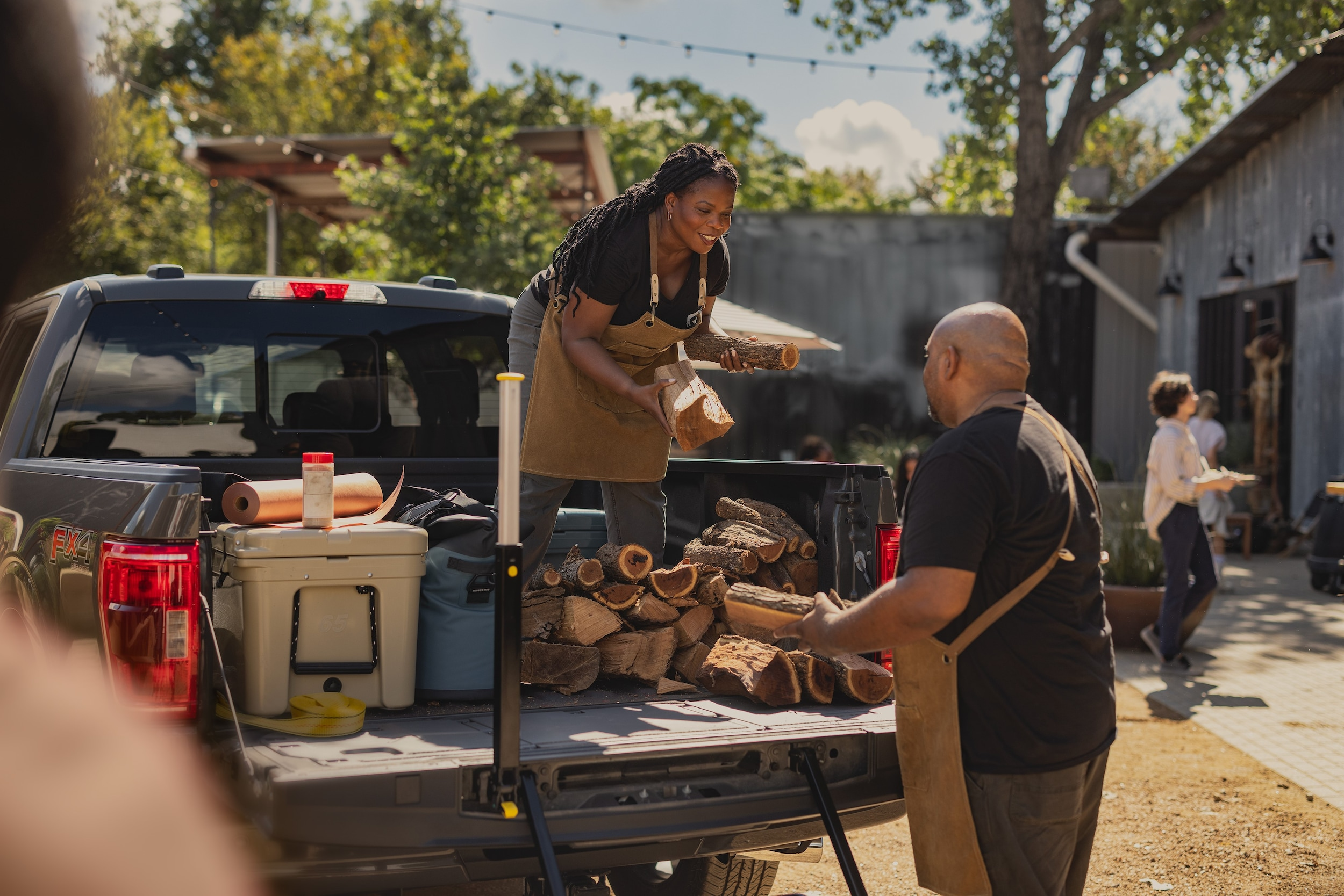 A man and woman, both wearing aprons, load firewood from a pickup truck in a sunny outdoor setting. Trees and people in the background create a lively atmosphere.