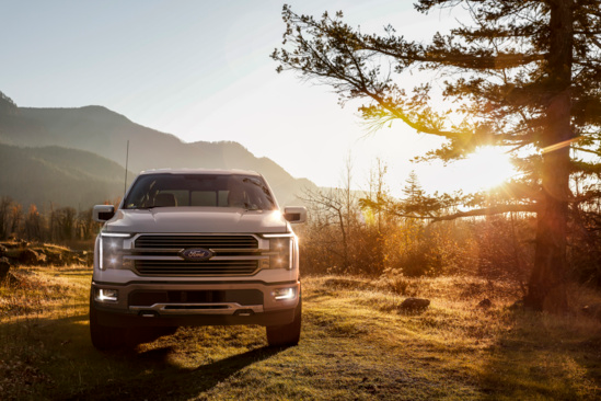 A white pickup truck is parked on a grassy field during sunset, surrounded by trees and mountains. The scene conveys a serene, adventurous vibe.